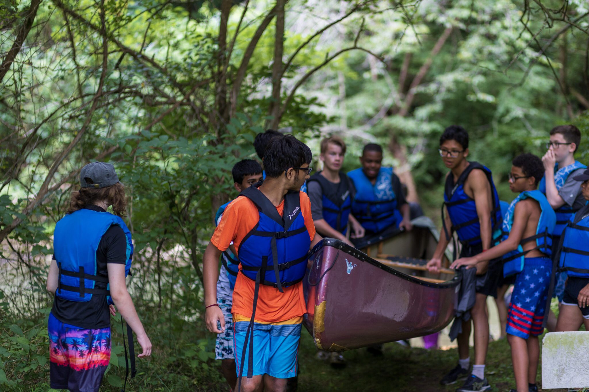 A group of people participating in the P.L.E.D.G.E. Leadership Program venture into the woods with their canoes.