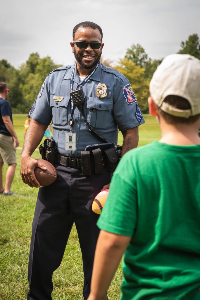 A police officer engaging with a young boy in a field as part of a Community Athletic Program.