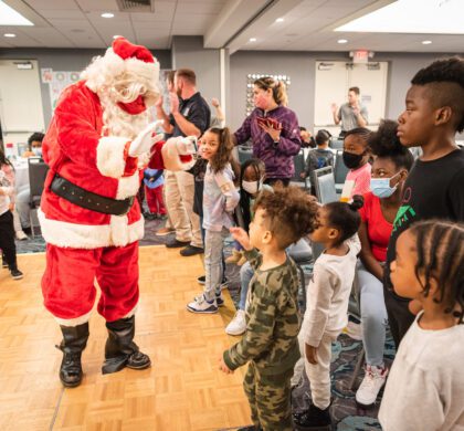 Santa Claus is standing in front of a group of children at the HoCo Kids Holiday Party.