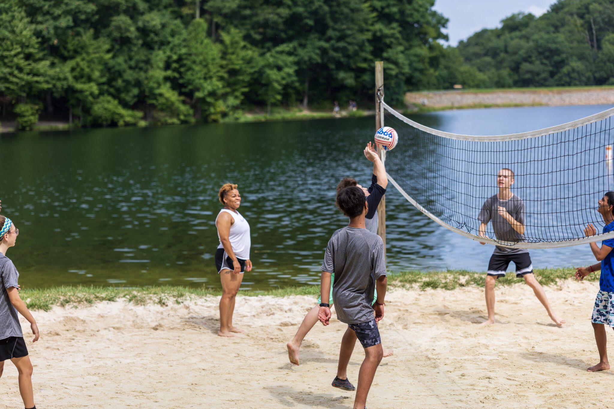 A group of people participating in the P.L.E.D.G.E. Leadership Program playing volleyball near a lake.