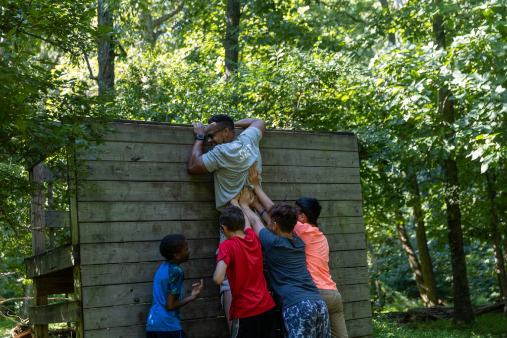 A group of adventurous kids embracing their BearTrax spirit as they fearlessly climb a wooden wall amidst the enchanting woods.