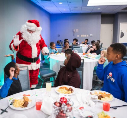 A Santa Claus is standing at a table with children during the HoCo Kids Holiday Party.