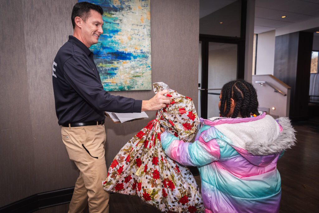 A man handing a girl a dress at the HoCo Kids Holiday Party.