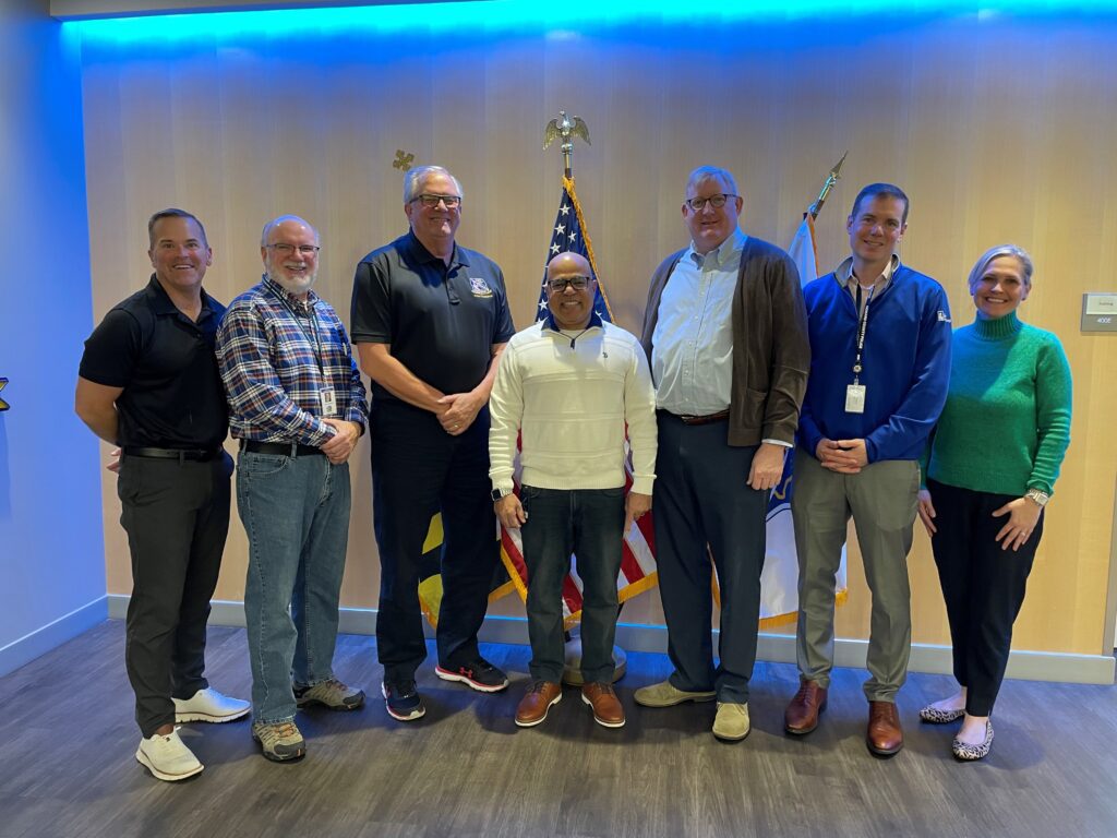 Group of six men standing indoors under blue lighting with an American flag in the background.